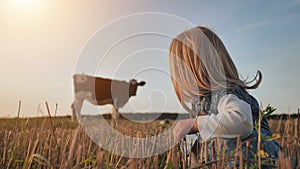 A little girl sits in a field against the backdrop of a lonely cow.