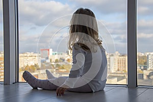 A little girl sits on the floor of an apartment in front of huge Windows and looks down at the city. Concept: children`s lonelines