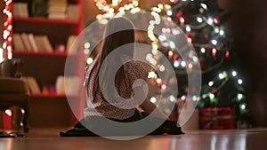 A little girl sits on the floor in the room in the evening and admires the Christmas tree.