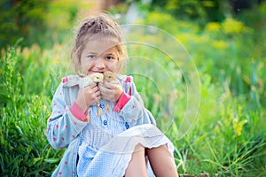 The little girl sits in a grass in the village and holds in hand of two chickens