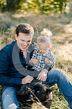 Little girl sits on the lap of her dad stroking the dog on the lawn