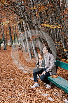 Little girl sits on the lap of a smiling dad on a bench in the autumn forest