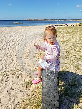A little girl sits near the sea on a tire buried in the ground until half