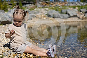 A little girl sits on a pebble and wets her feet in the water. A child on the shore of a pond carefully examines the