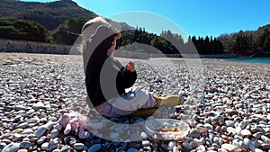 Little girl sits on a pebbly beach and eats porridge from a lunch box with a spoon