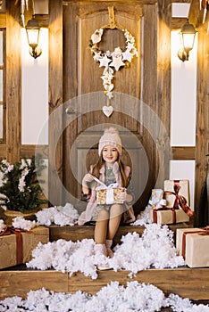 A little girl sits on the porch of a house decorated for Christmas and New Year. She holds a gift box in her hands, opens it by th