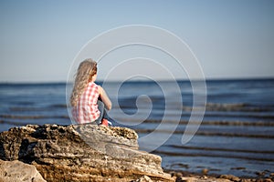 Little girl sits on a rocky shore and looks at the sea.