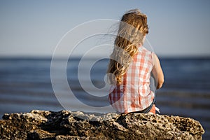 Little girl sits on a rocky shore and looks at the sea.