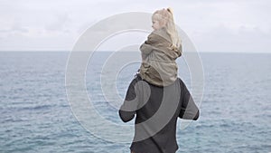 A little girl sits on the shoulders of her father and looks at the sea