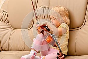 A little girl sits on a sofa with a violin and does not know what to do with it
