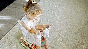 Little girl sits on stack of children's books and plays with her smartphone