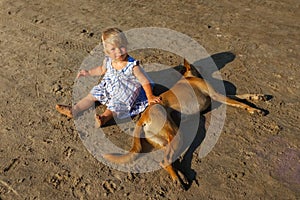 A little girl sits and strokes a red dog on the beach, the sun glints and color highlights.