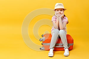 Little girl sits on a suitcase and holds cheeks with palms on a yellow background. A child is waiting for travel, trip, adventure