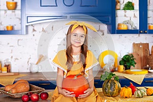 Little girl sits on table in kitchen holds big pumpkin. Harvesting. Healthy nutrition, vegetarianism, vitamins, vegetables. Cute b