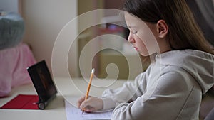 A little girl sits at the table and writes in a exercise book with a pencil. A tablet standing on the table with an