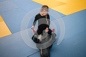 A little girl sits on top on a training mannequin and works off punches. Children`s sports activity in the gym. The child beats