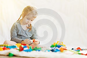 A little girl sits in a toy room on a white carpet and plays in a colored plastic constructor