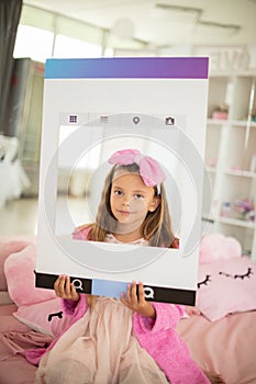 Little girl sitting on bed in bathrobe and lookin g at camera. Holding the frame photo