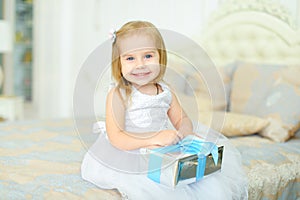 Little girl sitting on bed with present and wearing white dress.
