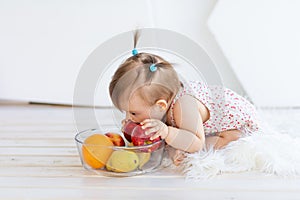 A little girl is sitting in a bright room with a plate of fruit and eating an Apple
