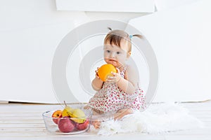 A little girl is sitting in a bright room with a plate of fruit and eating an orange