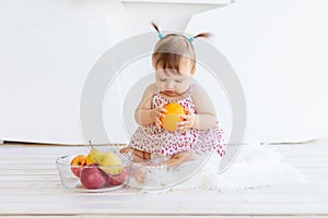 A little girl is sitting in a bright room with a plate of fruit and eating an orange