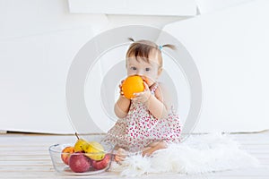 A little girl is sitting in a bright room with a plate of fruit and eating an orange