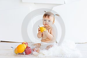 A little girl is sitting in a bright room with a plate of fruit and eating a pear