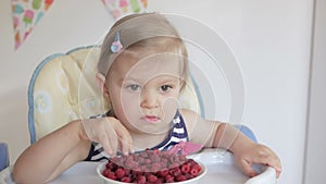 A little girl sitting at a children`s table eats ripe red raspberries