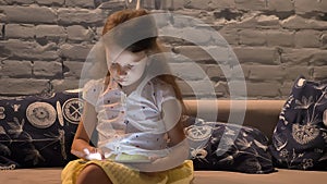 Little girl sitting on couch and typing on tablet, kid playing on gadget, child at home concept, indoors