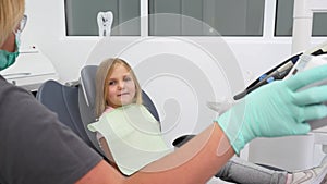 Little girl sitting in dentist's chair in modern dental office. Female dentist adjusting chair with child