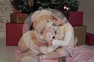 Little girl sitting on the floor with two different sizes toy bears waiting for Santa on Christmas Eve