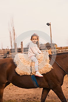 Little girl, sitting on a fluffy saddle and riding a brown-furred horse at a stable, outdoors
