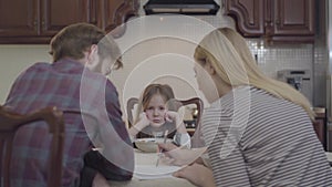 Little girl sitting in front of parents sad and unhappy, she does not eat. Young woman and man sitting at the table