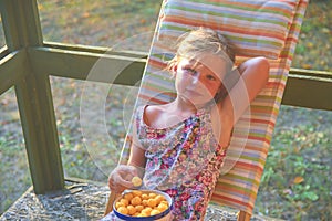 The little girl is sitting on a garden deckchair on a verandah. Small girl is eating cheese flavoured snacks in the