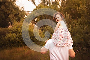 Little girl is sitting on her father`s shoulders in the summer in the park