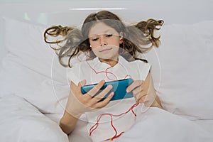 Little girl sitting at home in bed, with smartphone and headphones