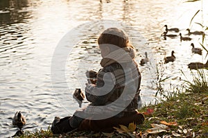 A little girl sitting on a lake side, looking at the water and feeding ducks