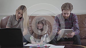 Little girl sitting between mother and father drawing with colored markers. Young woman typing on her laptop talking by