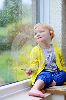 Little girl sitting next window on rainy day