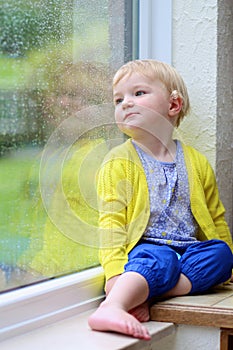Little girl sitting next window on rainy day