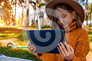 Little Girl Sitting In Park On Sunny Day And Play With Tablet Pc In Autumn Park.