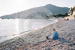 Little girl is sitting on a pebbly beach by the sparkling sea. Side view