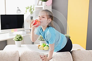 Little girl sitting on sofa and eating red apple at home