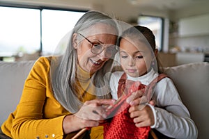 Little girl sitting on sofa with her grandmother and learning to knit indoors at home.