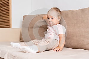 Little girl sitting on sofa and looking away with interest. Blond blue-eyed baby in light clothes