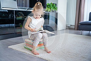 Little girl is sitting on a stack of children& x27;s books and leafing through a book with fairy tales