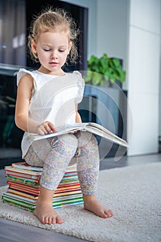 Little girl is sitting on stack of children& x27;s books and leafing through a book with fairy tales in home living room