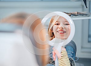 Little girl sitting in stomatology clinic chair and smiling at mirror showing her teeth after teeth dental procedures. Healthcare