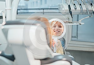 Little girl sitting in stomatology clinic chair and smiling at mirror showing her teeth after teeth dental procedures. Healthcare